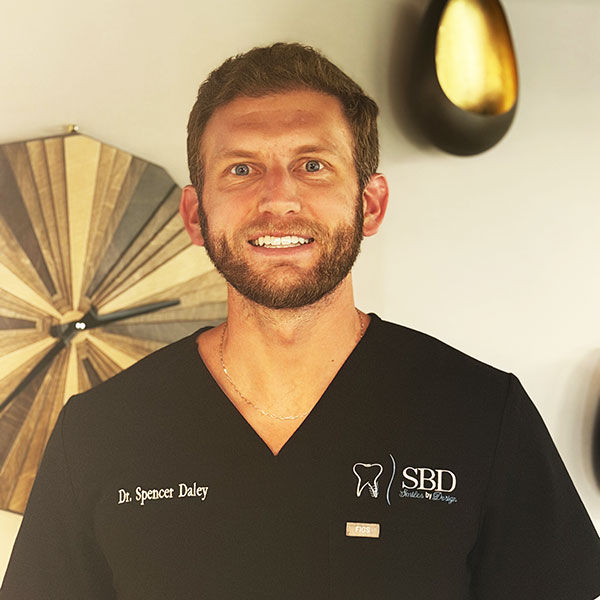 Dr. Spencer Daley in a black scrub top with SBD logo, smiling in a warmly lit room.