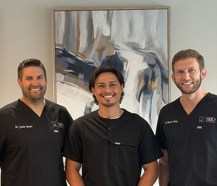 Three healthcare professionals in black scrubs pose for a group photo indoors, with a large abstract painting behind them.